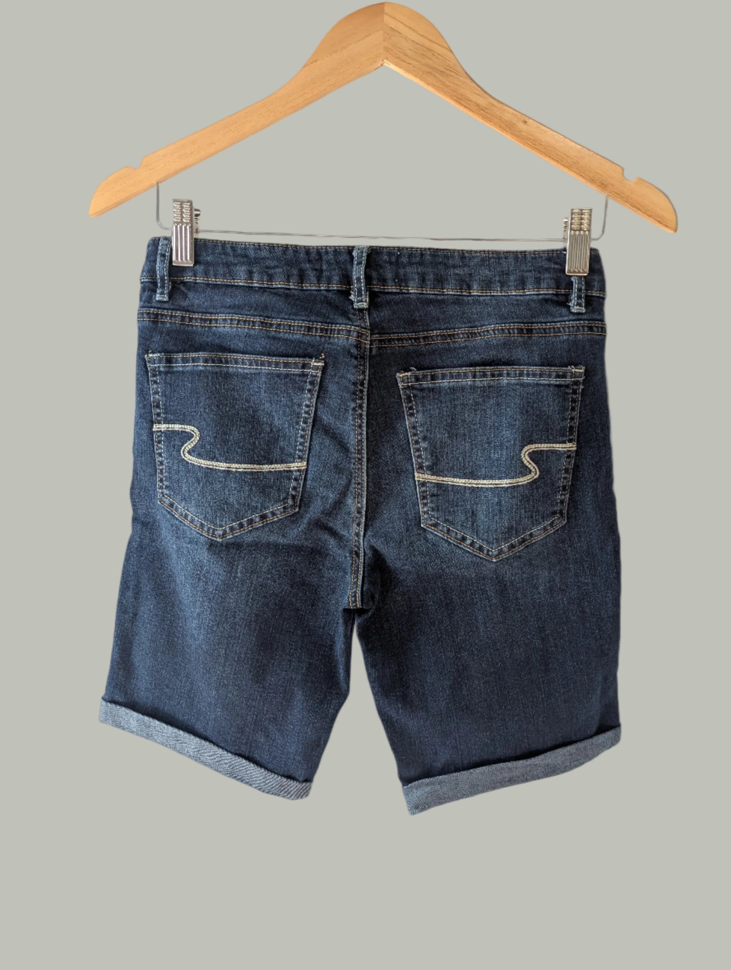 A back view of Eightytwo Dark Denim Mid-Thigh Shorts (Size 5) from Feather & Fable Thrift Co. with rolled cuffs hangs on a wooden hanger, shown from the back against a plain gray background.