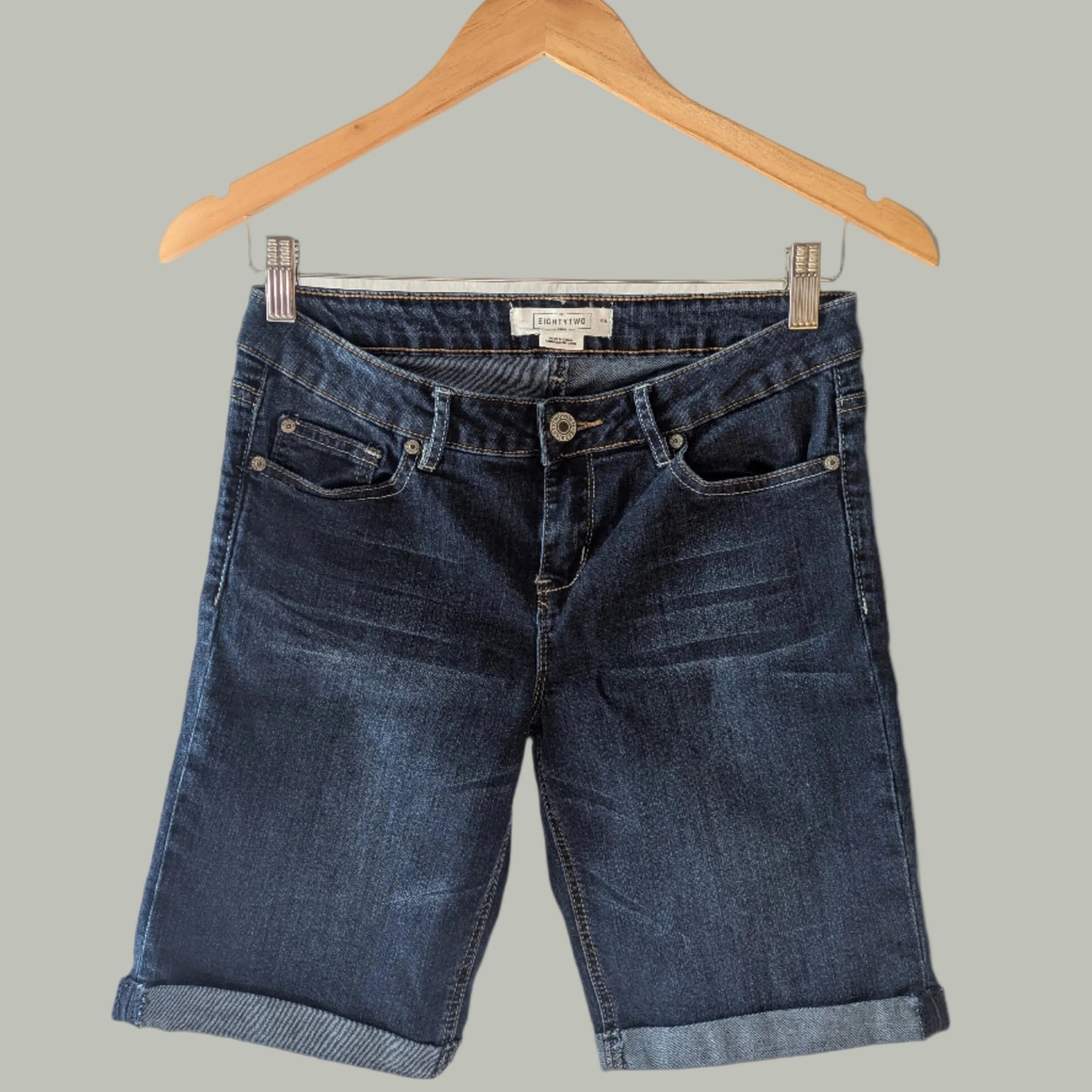 The Eightytwo Dark Denim Mid-Thigh Shorts (Size 5) by Feather & Fable Thrift Co. hang on a wooden hanger against a gray background, showcasing their stylish rolled-up cuffs.