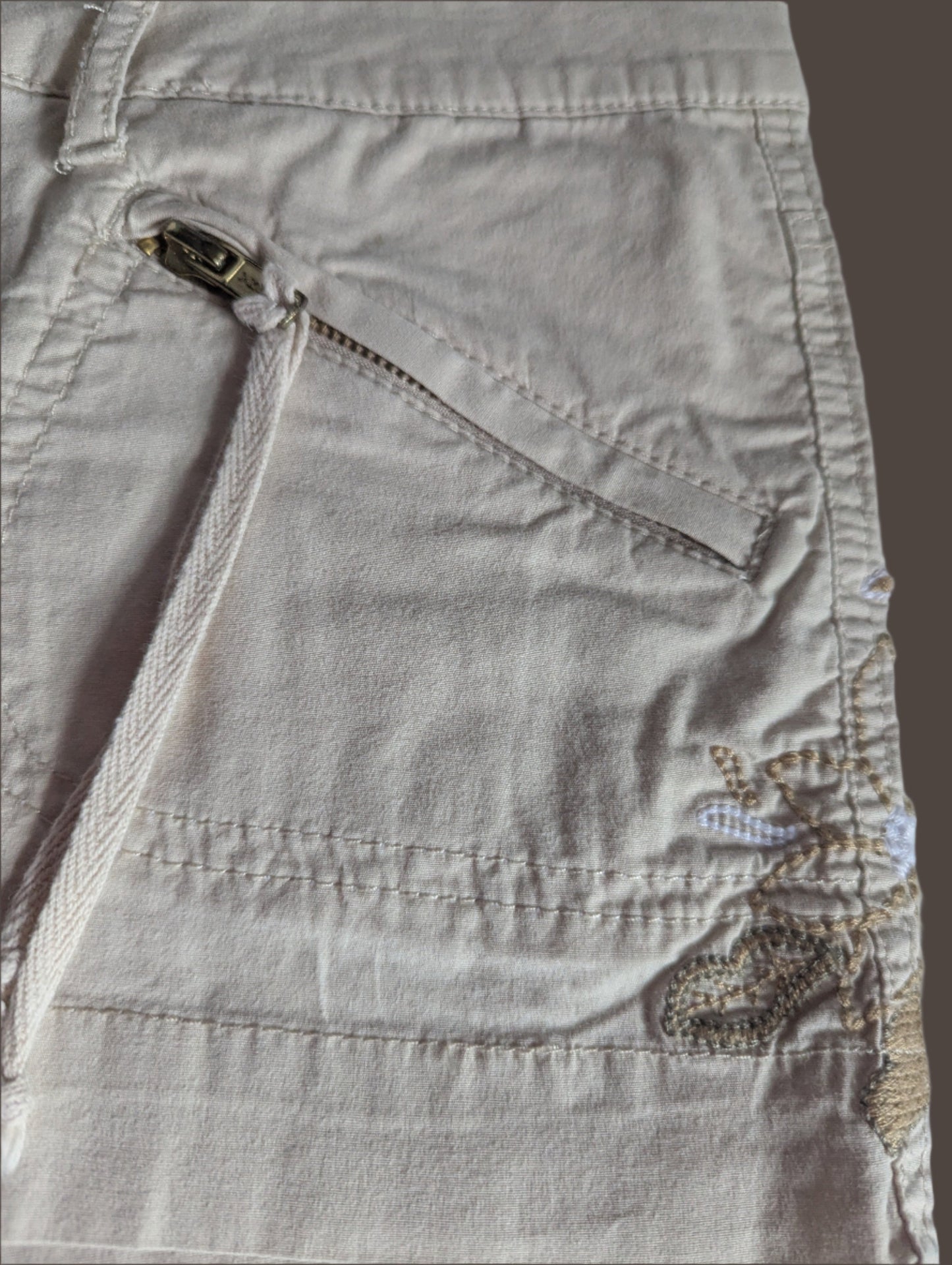 Close-up detail of the intricate embroidery along the waistband and the functional silver front zipper pocket on the Guess shorts.