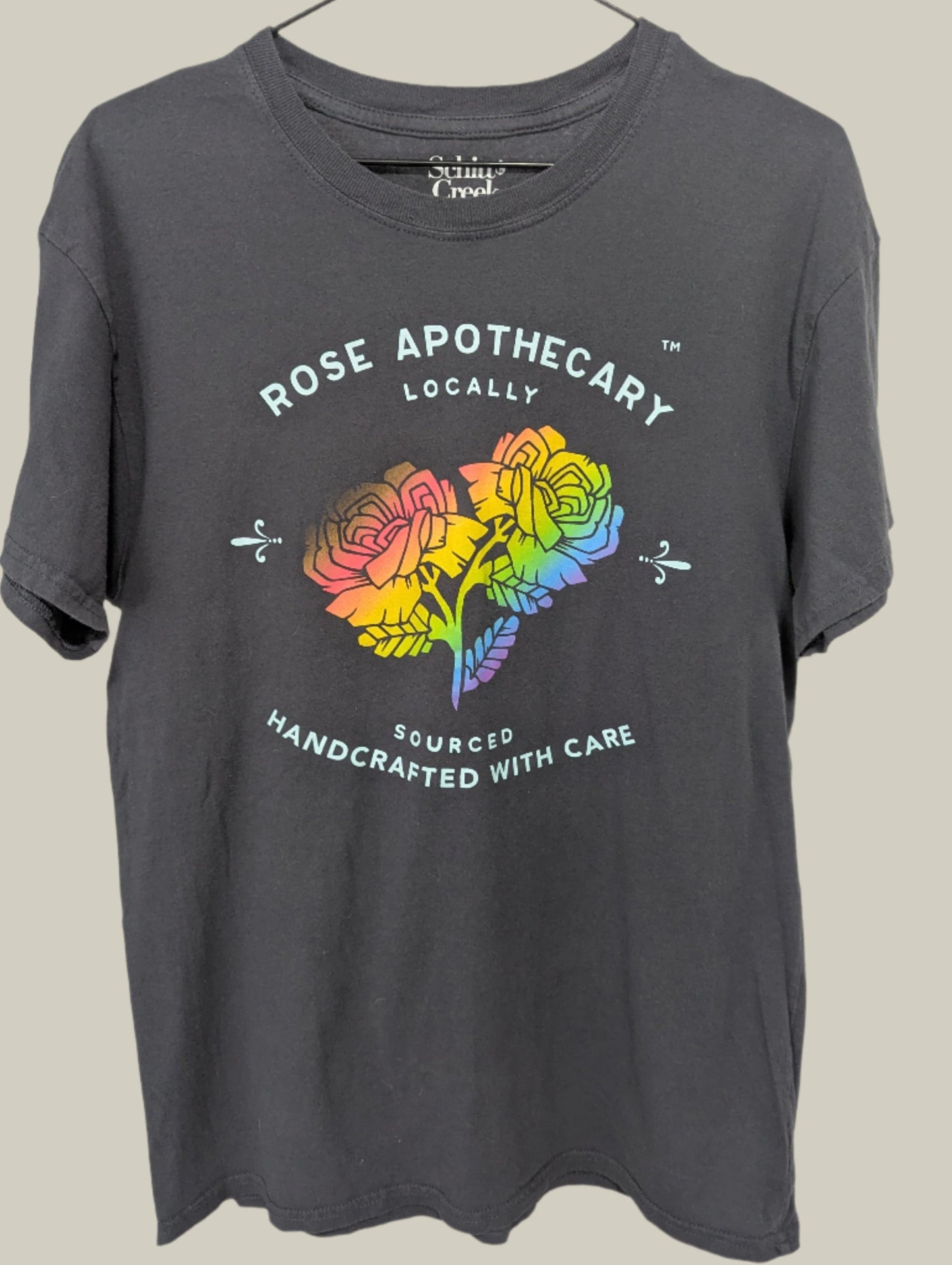 Front view of a unisex grey Schitt's Creek Rose Apothecary graphic t-shirt with a rainbow flower design hanging on a hanger.
