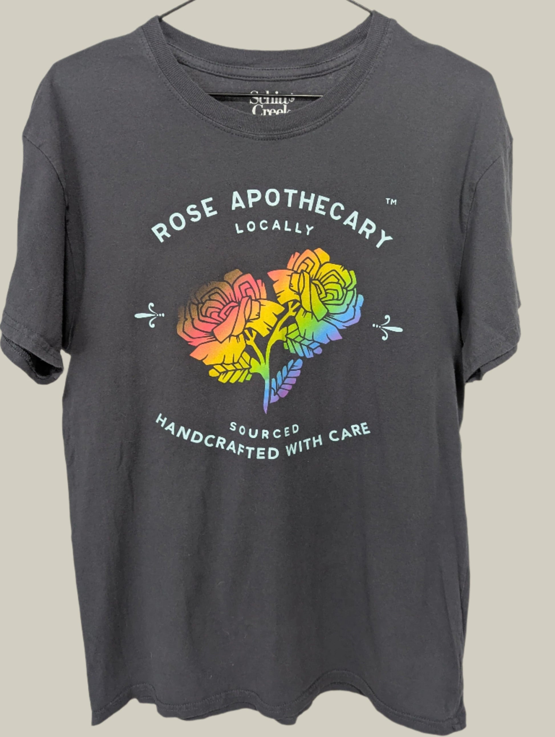 Front view of a unisex grey Schitt's Creek Rose Apothecary graphic t-shirt with a rainbow flower design hanging on a hanger.