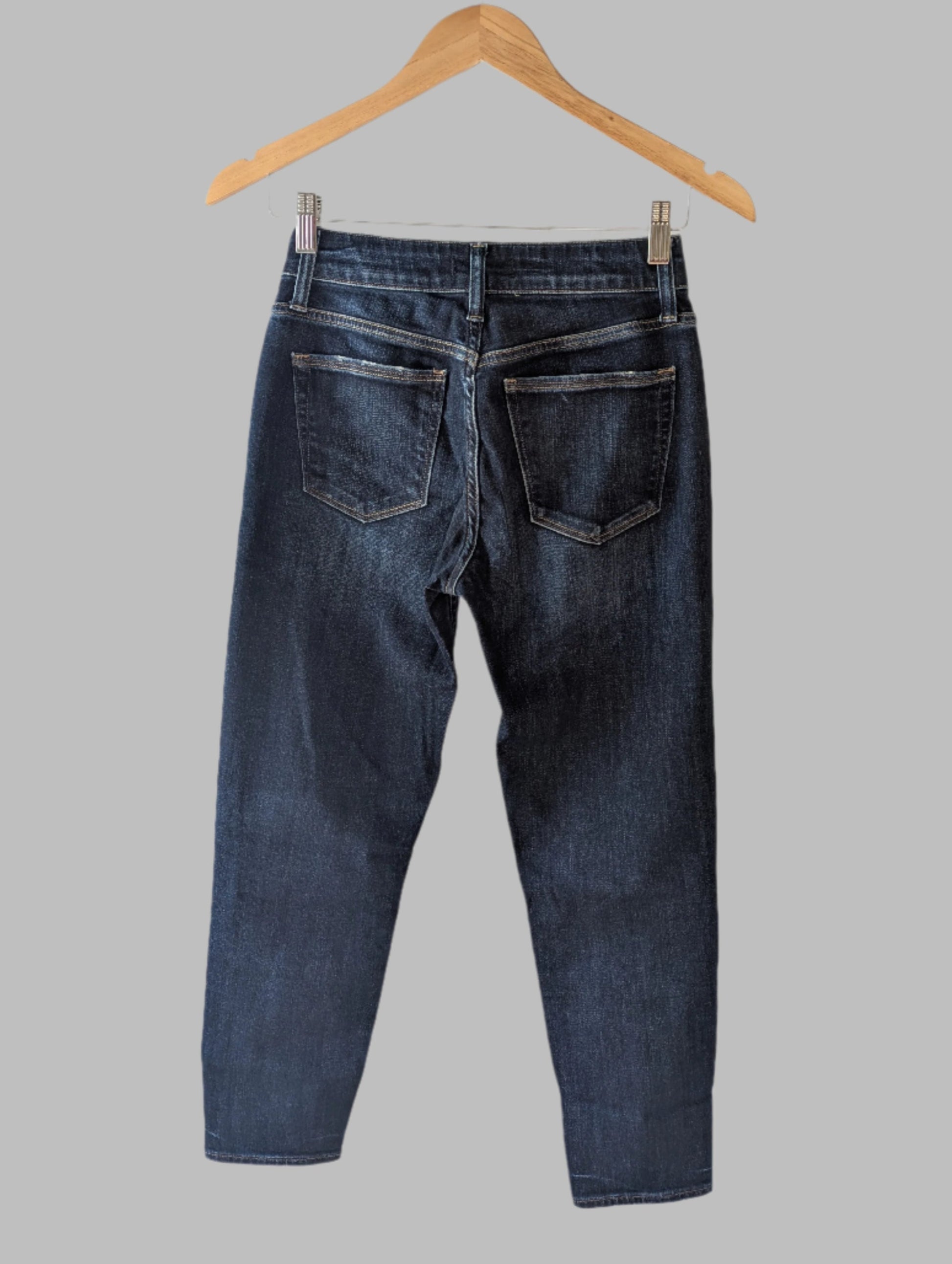 The Silver Jeans Avery Skinny Crop Dark Denim in size 25 by Feather & Fable Thrift Co. hangs on a wooden hanger, viewed from the back against a plain gray background.